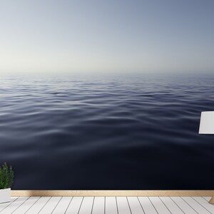 May include: A white lamp with a white lampshade sits on a white wood floor in front of a wall with a photo of a calm blue ocean.