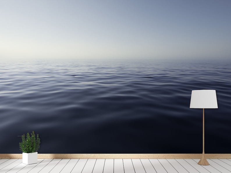 May include: A white lamp with a white lampshade sits on a white wood floor in front of a wall with a photo of a calm blue ocean.