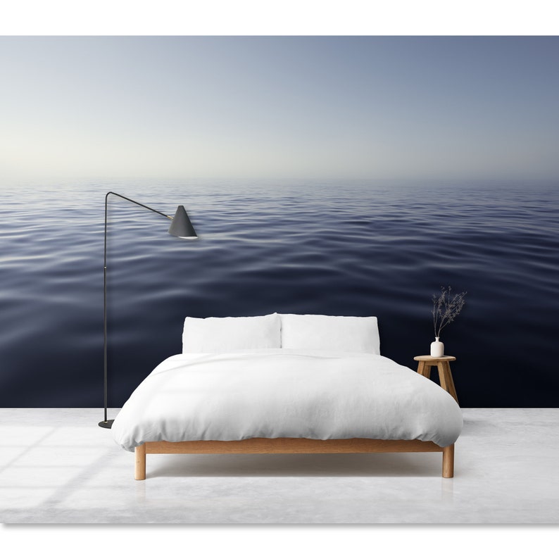 May include: A white bed with a white duvet and two white pillows sits in front of a wall mural depicting a calm, blue ocean.