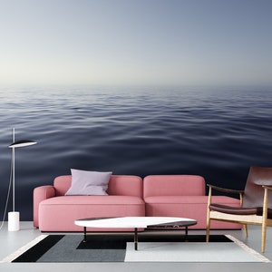 May include: A pink sofa with a purple pillow sits in front of a wall mural depicting a calm, blue ocean. A brown leather armchair and a white coffee table with a black marble top are also in the room.