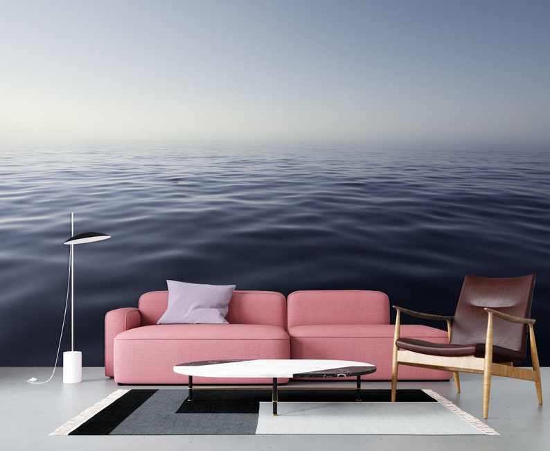 May include: A pink sofa with a purple pillow sits in front of a wall mural depicting a calm, blue ocean. A brown leather armchair and a white coffee table with a black marble top are also in the room.
