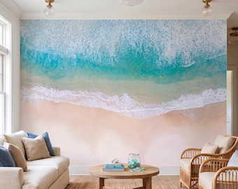 May include: A large wall mural featuring an aerial view of a sandy beach with turquoise ocean water and white sea foam waves. The mural covers the entire wall behind a beige sofa, a round wooden coffee table, and two rattan chairs.