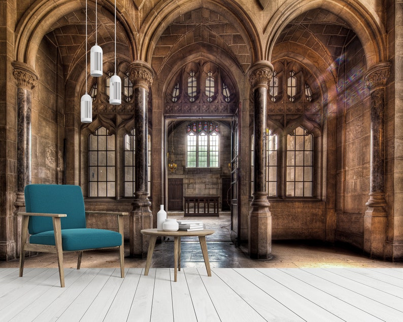 May include: An interior shot of a grand, stone-walled room with arched ceilings and large windows. A teal armchair and a small wooden table with books and vases are in the foreground. Overhead, three white pendant lights hang from the ceiling.