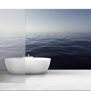May include: A white bathtub sits in a bathroom with a large window overlooking a calm, blue ocean.