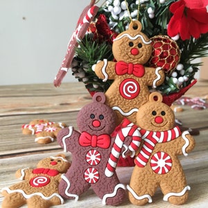 May include: Four gingerbread man ornaments with red and white candy cane accents. The ornaments are brown, tan, and dark brown. The ornaments are hanging from a Christmas tree.