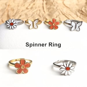 May include: Five gold and silver spinner rings with different flower and butterfly designs. The rings are on a brown background. The text "Spinner Ring" is below the rings.
