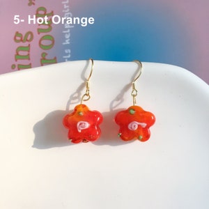 May include: A pair of gold dangle earrings with star-shaped glass beads. The beads are orange, yellow, and pink with green and white accents. The earrings are labelled "5- Hot Orange".