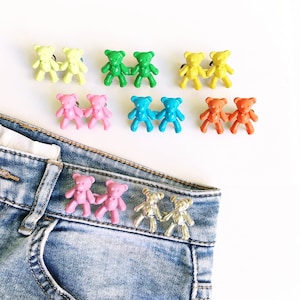 May include: A set of 10 pairs of colourful bear-shaped buttons. The buttons are in various colours, including yellow, green, blue, pink, and orange. The buttons are arranged in pairs, with each pair facing each other. The buttons are attached to a pair of blue jeans.