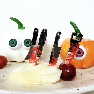 Puede incluir: Cuatro cuchillos en miniatura con detalles rojos y negros, utilizados como pinchos para comida, se insertan en una calabaza blanca, una calabaza naranja y una fruta blanca.