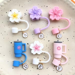 Pode incluir: Seis rolhas de garrafa de silicone coloridas em forma de flores de cerejeira, com um pequeno amuleto preso a cada uma. Os amuletos são dourados e têm as letras A a F.