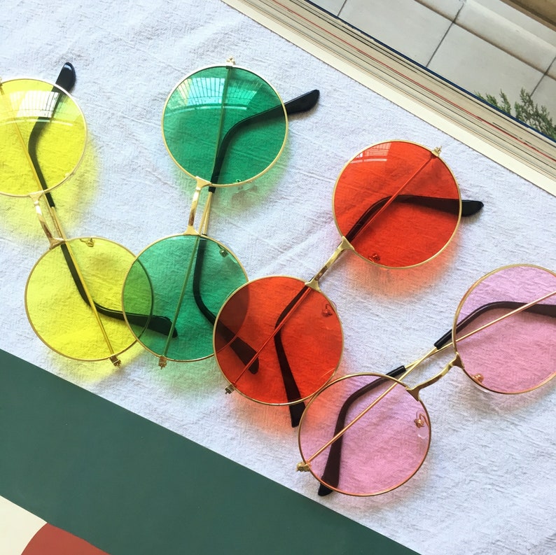 May include: Five pairs of round sunglasses with different colored lenses. The lenses are yellow, green, red, pink, and blue. The frames are gold.