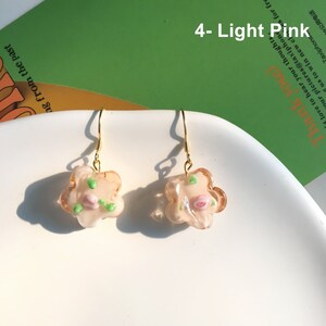 May include: A pair of gold dangle earrings with a light pink flower charm. The flower charm has a pink rose and green leaves.