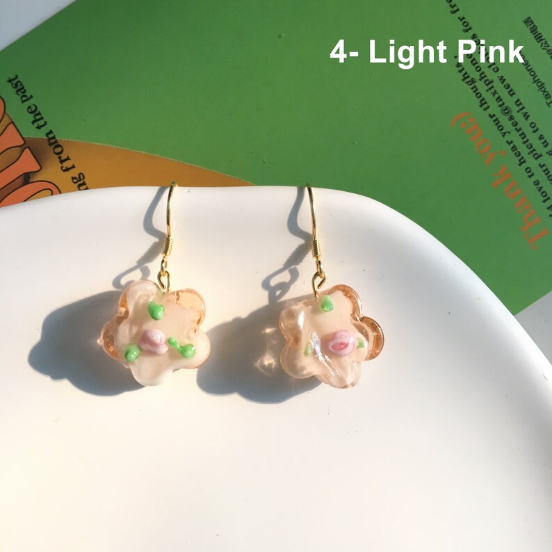 May include: A pair of gold dangle earrings with a light pink flower charm. The flower charm has a pink rose and green leaves.
