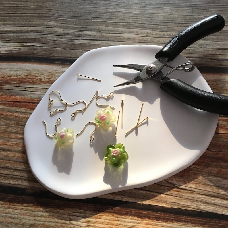 May include: A white plate with a pair of pliers, earring hooks, and small glass flower charms in green and white. The charms have pink flower details.