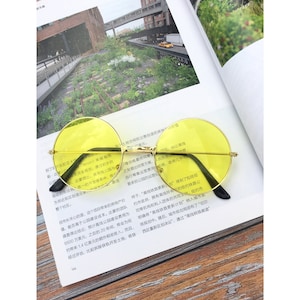 May include: Round yellow-tinted sunglasses with a gold metal frame. The sunglasses are lying on a page of a book.