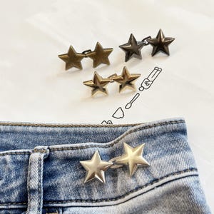 May include: Four pairs of star-shaped metal waist adjusters in various metallic finishes including antique brass, dark gunmetal, polished gold, and silver. One silver pair is attached to the waistband of light blue denim jeans.