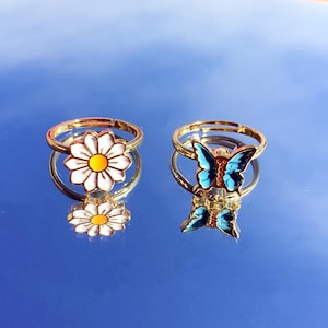 Puede incluir: Dos anillos de oro con diseños de esmalte. Un anillo presenta una margarita blanca con un centro amarillo. El otro anillo presenta una mariposa azul con acentos naranjas.