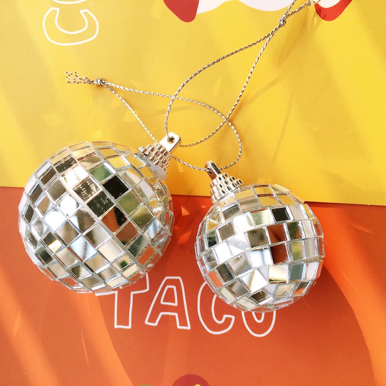 May include: Two silver disco ball ornaments hanging from a string. The ornaments are made of small mirrored tiles and are hanging against a yellow and orange background.