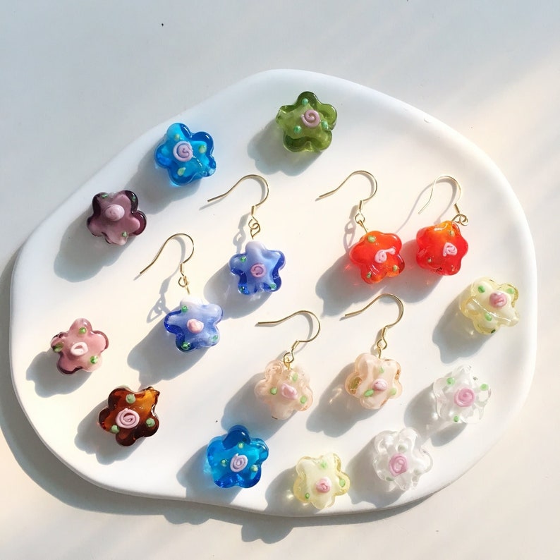 May include: Assortment of colourful glass flower earrings with gold hooks. The earrings feature a variety of colours, including blue, green, red, pink, and orange. Each earring has a small flower design with a pink centre.