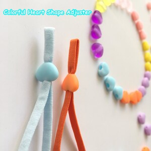 May include: Two heart-shaped adjustable cord stoppers, one light blue and one orange, on a white background. The stoppers are attached to elastic cords, one light blue and one orange. The text "Colorful Heart Shape Adjuster" is written in blue at the top of the image.