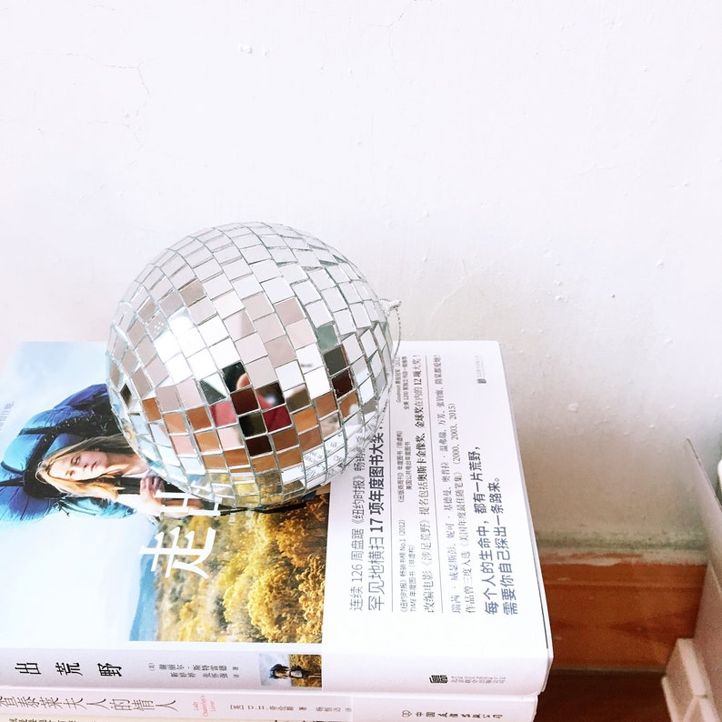 May include: A silver disco ball sits on top of a stack of books. The books are in Chinese and English. The text on the books reads "Out of the Wilderness" and "Lady Chatterley's Lover".
