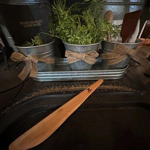 May include: Three galvanized metal pots with burlap bows and greenery are arranged on a metal tray. A wooden kitchen utensil with the word "TNT" is on a black surface.