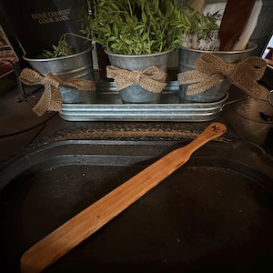 May include: Three galvanised metal planters with burlap bows, filled with green leafy plants, sit on a rustic wooden tray. A wooden spatula rests on a black cast iron pan.