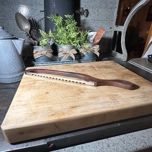 May include: A wooden cutting board with a serrated bread knife on top. The cutting board is on a gray surface with a gray enamel coffee pot and three small metal pots with green plants and burlap bows.