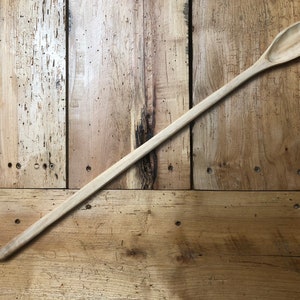 Puede incluir: Una cuchara de madera larga con una cabeza redonda de forma ovalada. La cuchara es de color marrón claro y está sobre una superficie de madera.