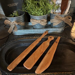 May include: Three wooden spatulas with a brown finish and a black engraved design. The spatulas are arranged on a black oval tray with a woven brown surface underneath.
