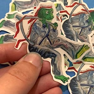May include: A die-cut sticker featuring a green frog dressed in a white sailor uniform with a red neckerchief. The frog is depicted in a seated pose, wearing a white sailor hat with a red band, and holding a long, thin, curved gray object.