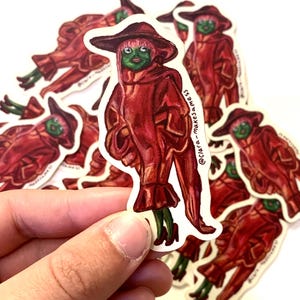 Peut inclure: Un autocollant découpé représentant un personnage à la peau verte et aux cheveux roses, portant un chapeau rouge à larges bords, un long manteau rouge à manches à volants et des chaussures rouges. Le texte @clara-makesamess est imprimé verticalement.