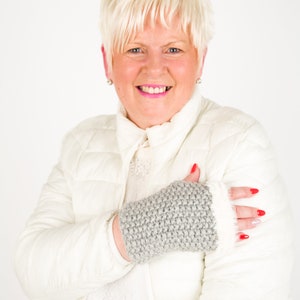 May include: A woman wearing a white puffer jacket with a gray knitted fingerless glove on her left hand. She is smiling and looking at the camera.