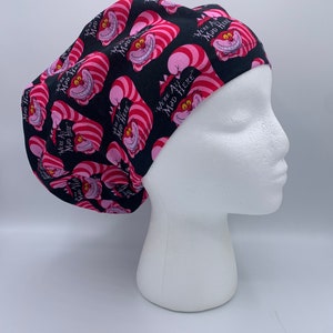 May include: A black scrub cap featuring a repeating pattern of the Cheshire Cat from Alice in Wonderland. The cat is pink and red with a wide grin. Text on the cap reads "We're All Mad Here".