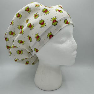 May include: A white scrub cap featuring a repeating pattern of green frog faces with red mouths and yellow collars. The fabric has a subtle yellow polka dot background. The cap is shown on a white mannequin head, displaying a rounded, gathered design.