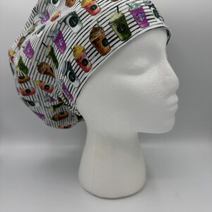May include: A white and black striped scrub cap featuring colorful coffee drinks. The cap is adorned with various coffee cups and beverages in shades of brown, green, pink, and white. The cap is displayed on a white mannequin head.