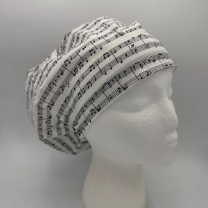 May include: A white scrub cap featuring a black musical staff and notes pattern. The cap is made of fabric and is designed to cover the head. The cap is displayed on a white mannequin head.