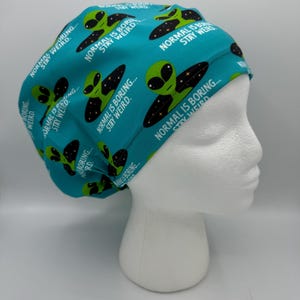 May include: Teal surgical scrub cap with a repeating pattern of green alien heads and the text "NORMAL IS BORING... STAY WEIRD." The cap is designed to fit comfortably and securely.