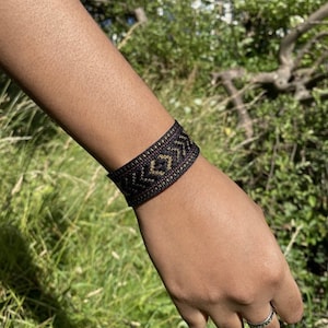 Puede incluir: Un brazalete tejido negro con un patrón geométrico en oro y rojo. El brazalete se lleva en la muñeca.