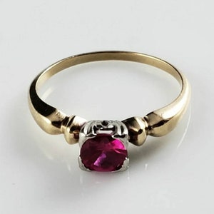 Puede incluir: Un anillo de oro con una piedra preciosa roja engastada en un metal blanco. El anillo tiene una banda simple con un diseño ligeramente elevado.