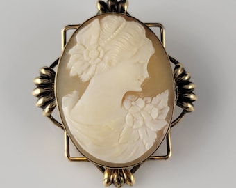May include: A gold-toned cameo brooch featuring a carved shell cameo of a woman's profile with floral details. The cameo is set in a square frame with ornate details.