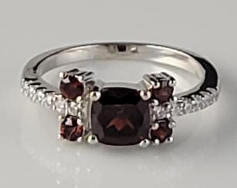 May include: A silver ring with a large, square-shaped, dark red gemstone surrounded by smaller red gemstones and clear cubic zirconia stones.
