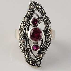 May include: A silver ring with a delicate, leaf-shaped design featuring three round, ruby-red gemstones and a cluster of marcasite stones.