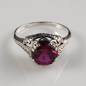 Könnte beinhalten: Ein silberner Ring mit einem ovalen, tief roten Edelstein, der in einem filigranen Design gefasst ist.