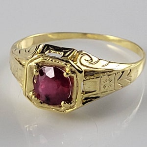 May include: A gold ring featuring a vibrant, deep red gemstone in a square setting. The ring's band is intricately engraved with patterns, and the overall design suggests a vintage aesthetic. The gemstone is the central element, set within the polished gold.