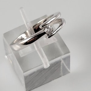 Puede incluir: Un anillo plateado con una sola gema transparente. El anillo tiene un diseño único y fluido con una banda dividida que se curva alrededor de la gema. El anillo se exhibe sobre un soporte rectangular transparente.