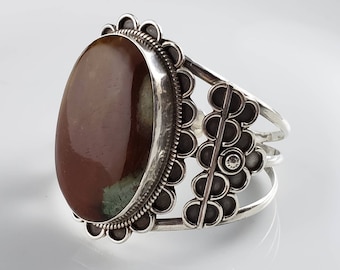May include: A silver cuff bracelet featuring a large oval-shaped brown and green gemstone set in the centre. The bracelet has a decorative design of small circles around the gemstone and a band of circles on the side.