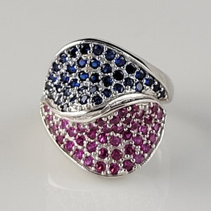 Puede incluir: Un anillo de plata con un diseño de piedras azules y rosas. El anillo tiene forma de hoja y presenta un patrón ondulado.