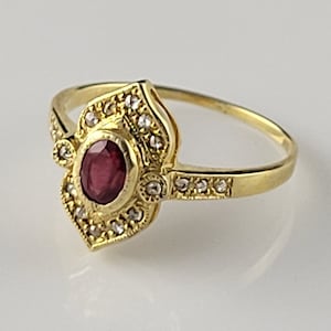 Op de afbeelding: Een gouden ring met een ovale, dieprode edelsteen in het midden, omringd door kleine, heldere stenen. De ring heeft een sierlijk filigraanontwerp met extra kleine stenen langs de band. De ring is geplaatst tegen een neutrale achtergrond.