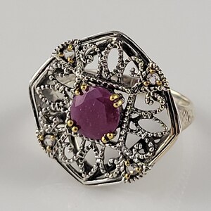 May include: A silver ring with a filigree design and a large, round, purple gemstone in the center. The ring is accented with small gold beads.